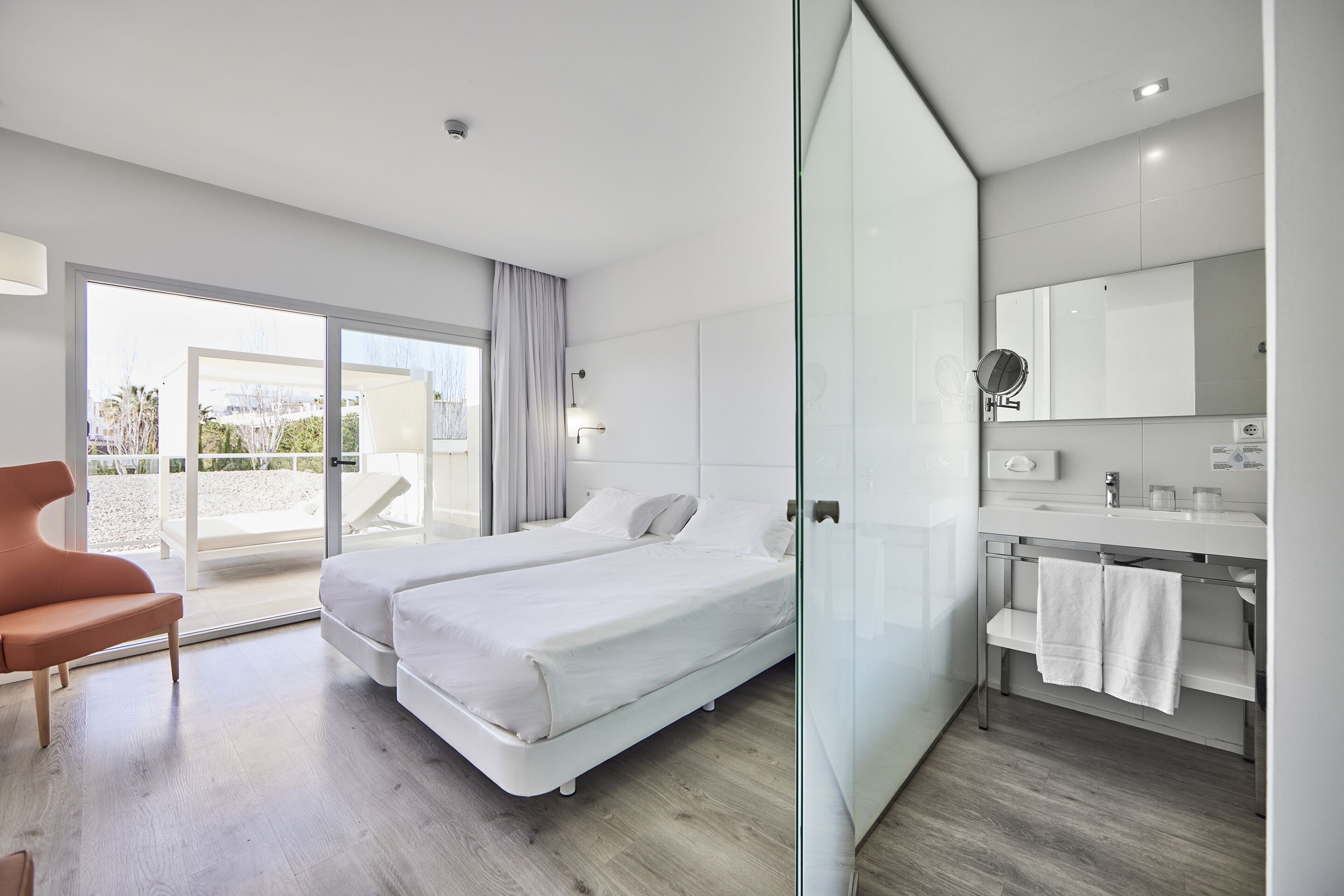 A modern hotel room with a large bed, a glass partition, a bathroom with a mirror, and a view of the outdoors.