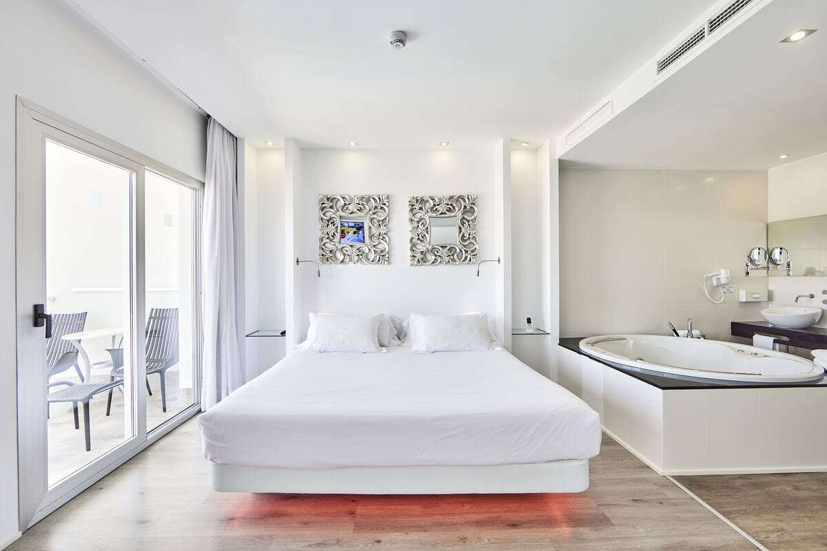Une chambre d’hôtel moderne dotée d’un grand lit, d’une salle de bain séparée avec baignoire, et d’une porte-fenêtre coulissante donnant sur un balcon avec coin salon extérieur.