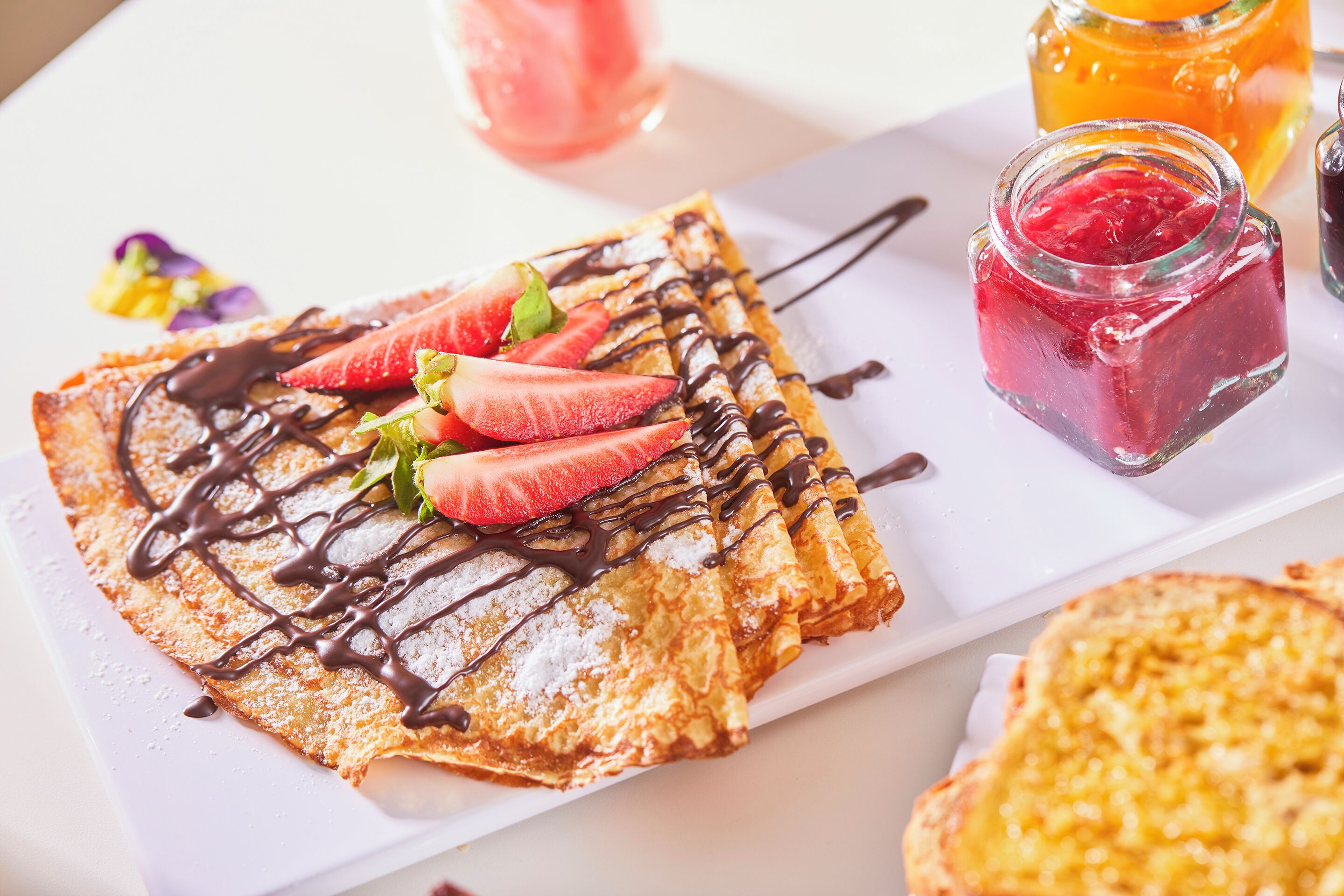 A plate of crepes with strawberries and chocolate sauce, accompanied by a jar of jam and a glass of juice.