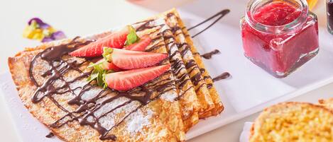 A plate of crepes with strawberries and chocolate sauce, accompanied by a jar of jam and a glass of juice.