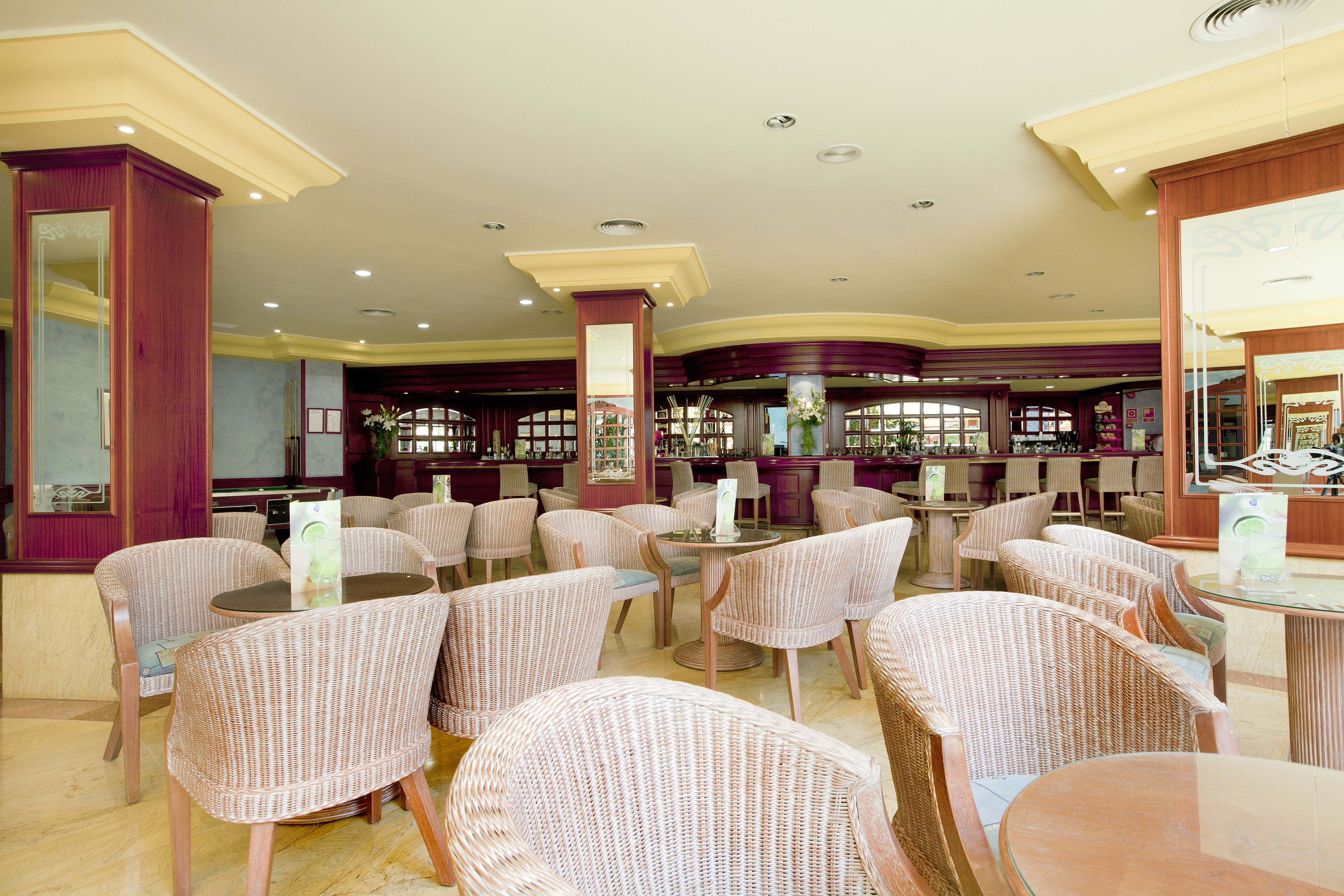 A spacious restaurant with wicker chairs and tables, a bar area, and a view through large windows.