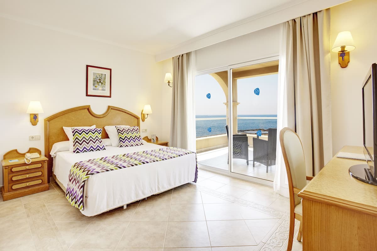 A hotel room with a bed, a desk, a chair, a TV, a lamp, a picture on the wall, and a view of the sea.
