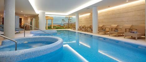 A modern indoor swimming pool with a mosaic tile design, lounge chairs, and a mural of ancient ruins.