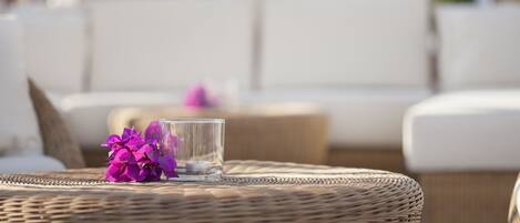 A wicker round table with a glass and purple flowers, set on a wicker ottoman.