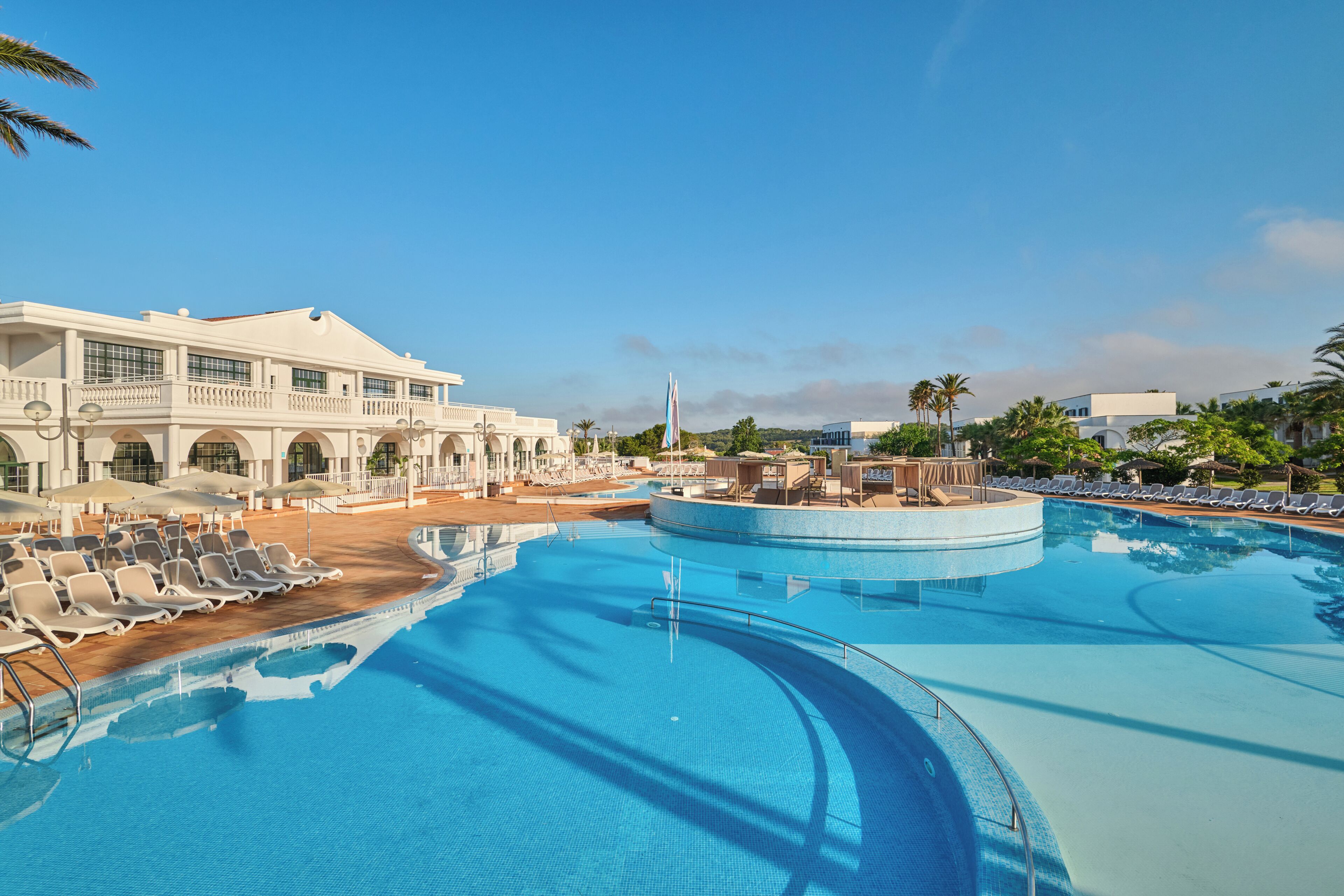 A large swimming pool with lounge chairs and umbrellas, surrounded by a building with a white facade and columns.