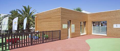 A building with a wooden exterior, a fenced playground area, and palm trees in the background.