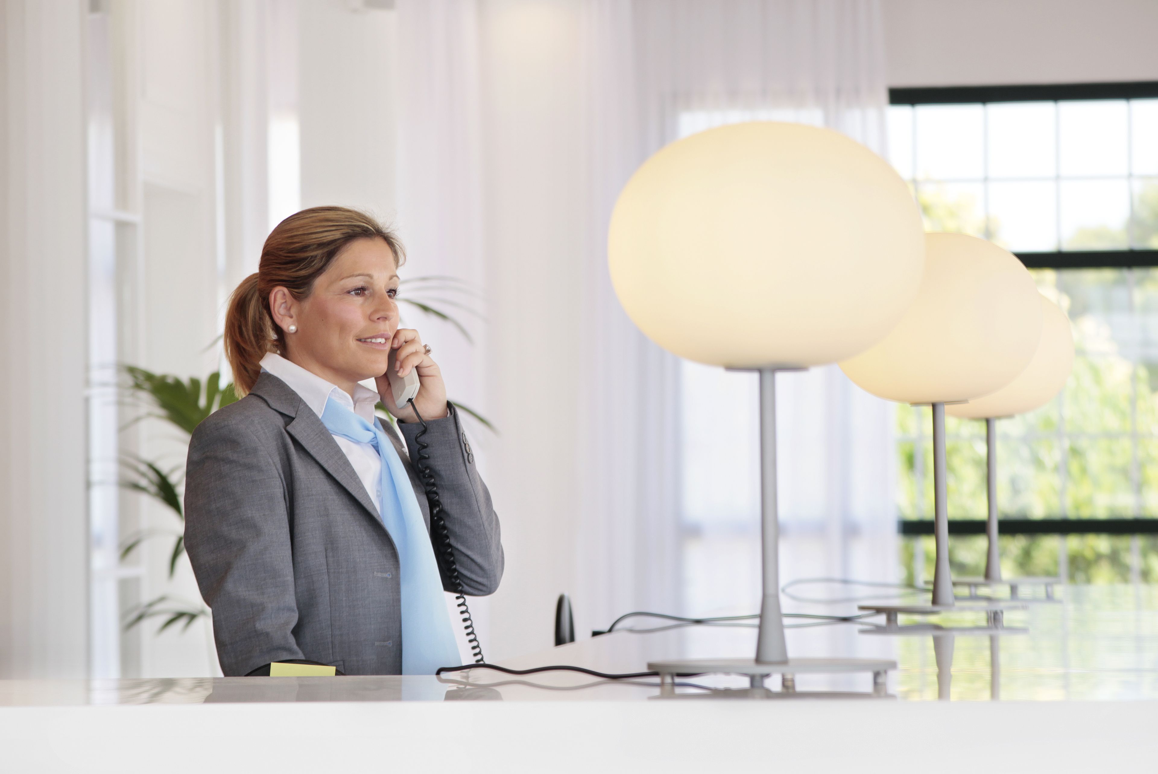 Een vrouw in een pak achter een bureau met een telefoon, in een modern kantoor met grote ramen en bolvormige lampen.