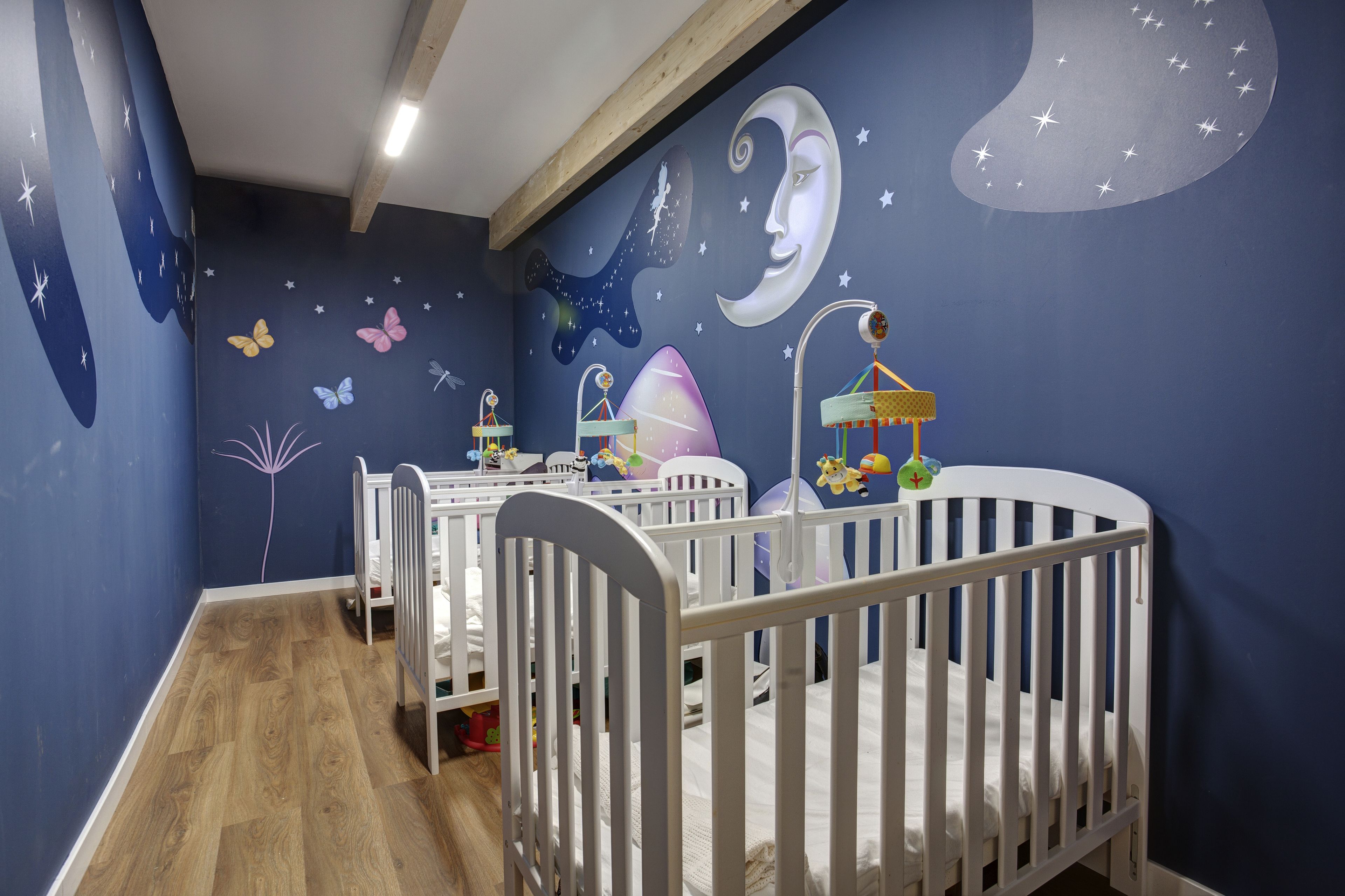Una habitación con dos cunas blancas, suelo de madera y un mural con estrellas, lunas y mariposas.