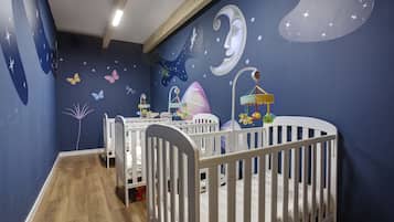 Una habitación con dos cunas blancas, suelo de madera y un mural con estrellas, lunas y mariposas.