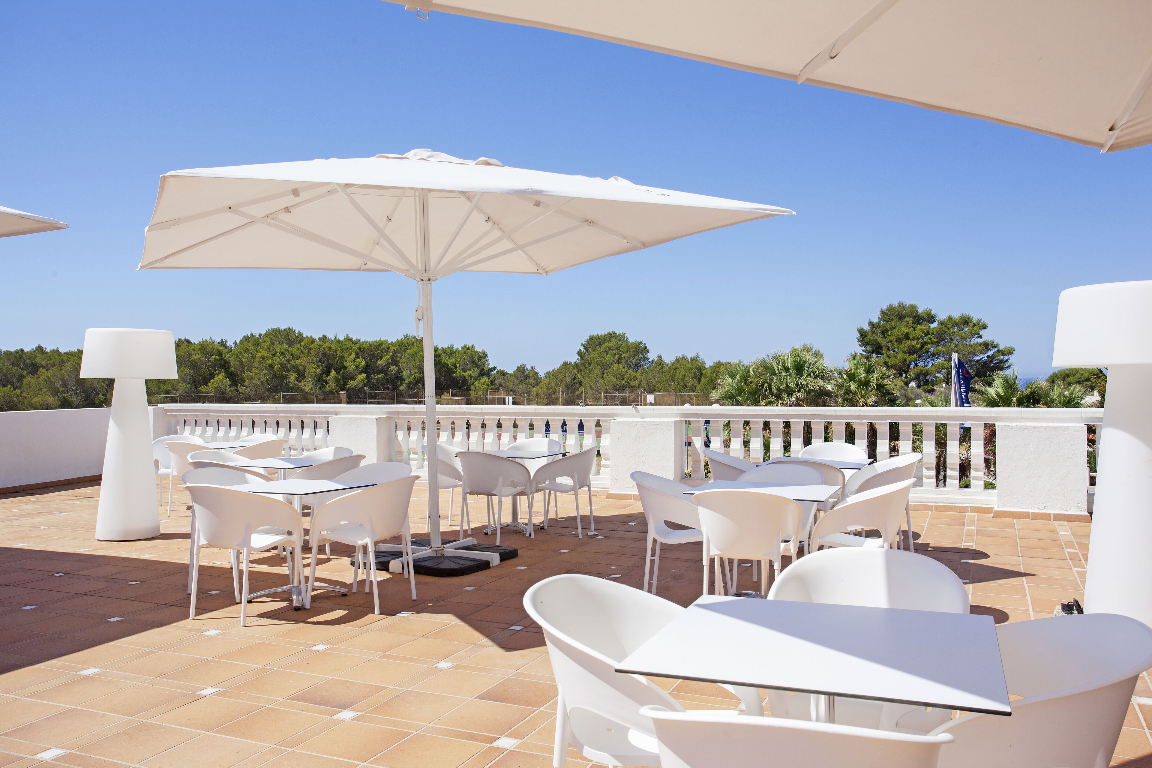 A terrace with white tables and chairs, large umbrellas, and a view of the sky and trees.