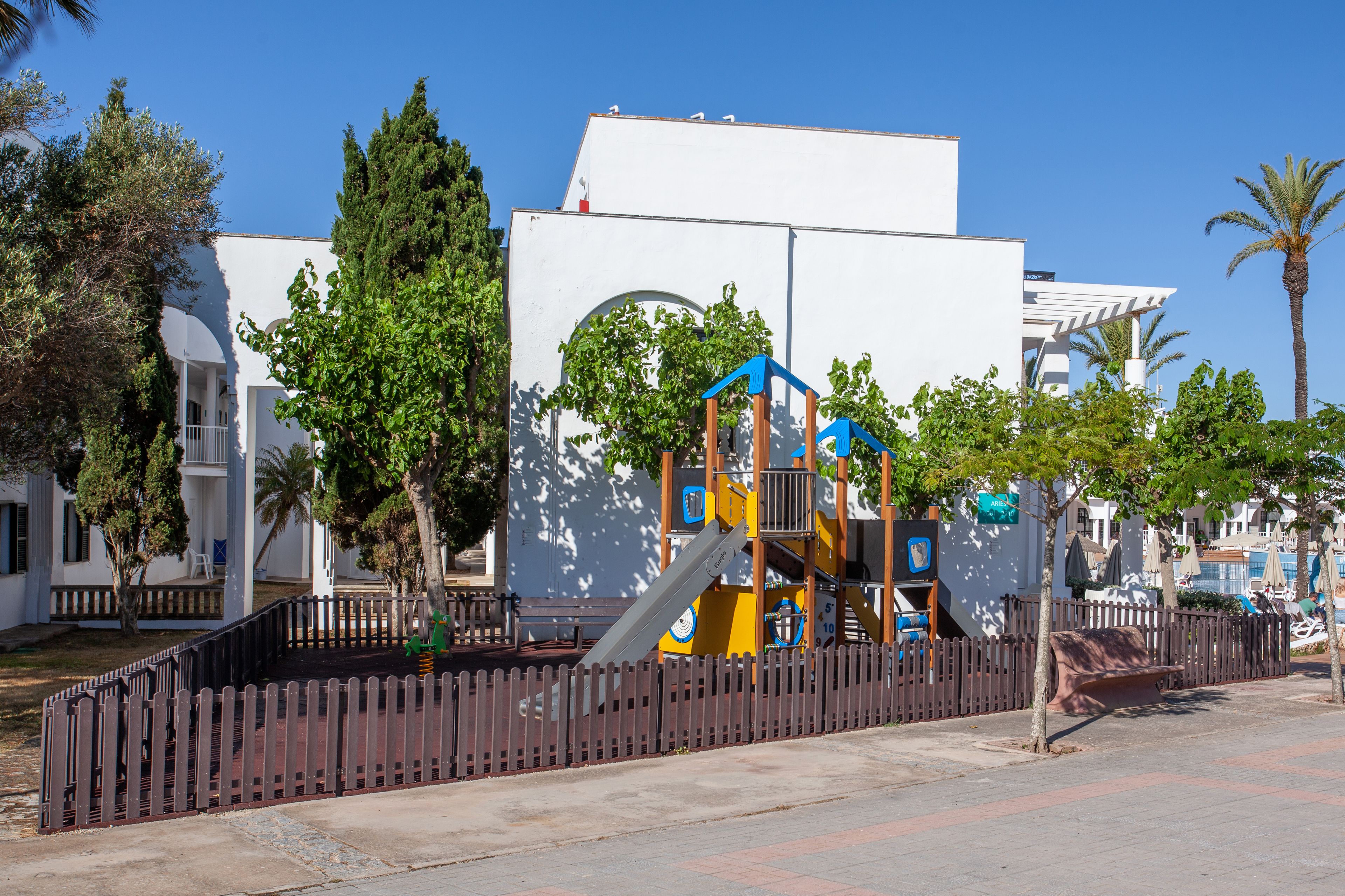 A playground with climbing structures and a fenced area, surrounded by trees and a white building with balconies.