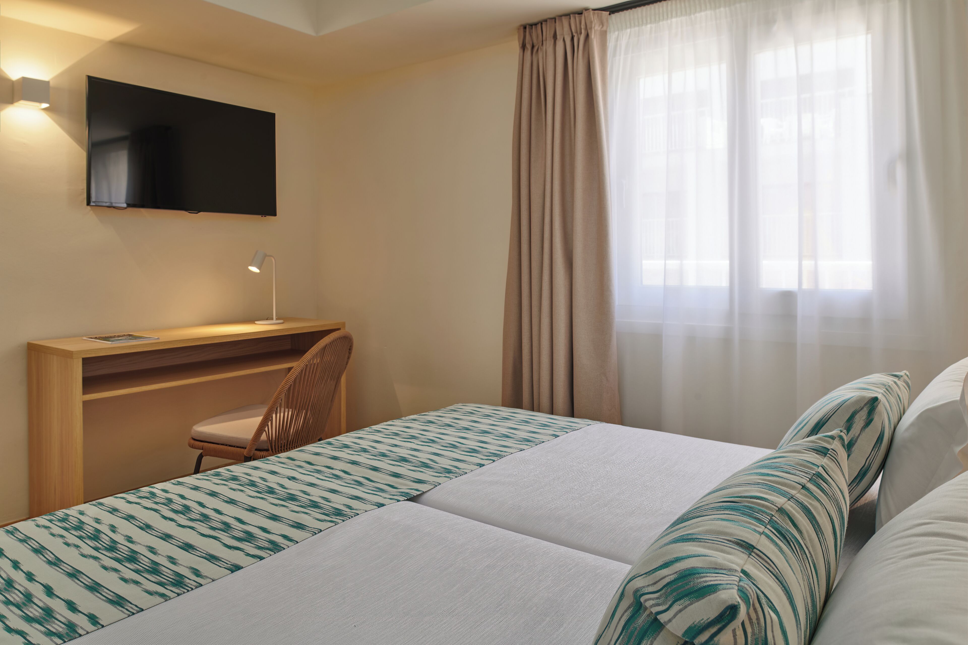Habitación de hotel con cama, escritorio, silla, televisor montado en la pared y ventana con cortinas.