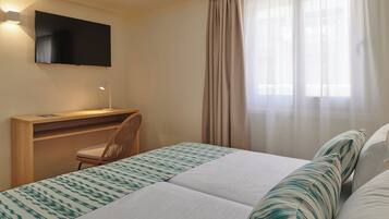 A hotel room with a bed, a desk, a chair, a TV mounted on the wall, and a window with curtains.
