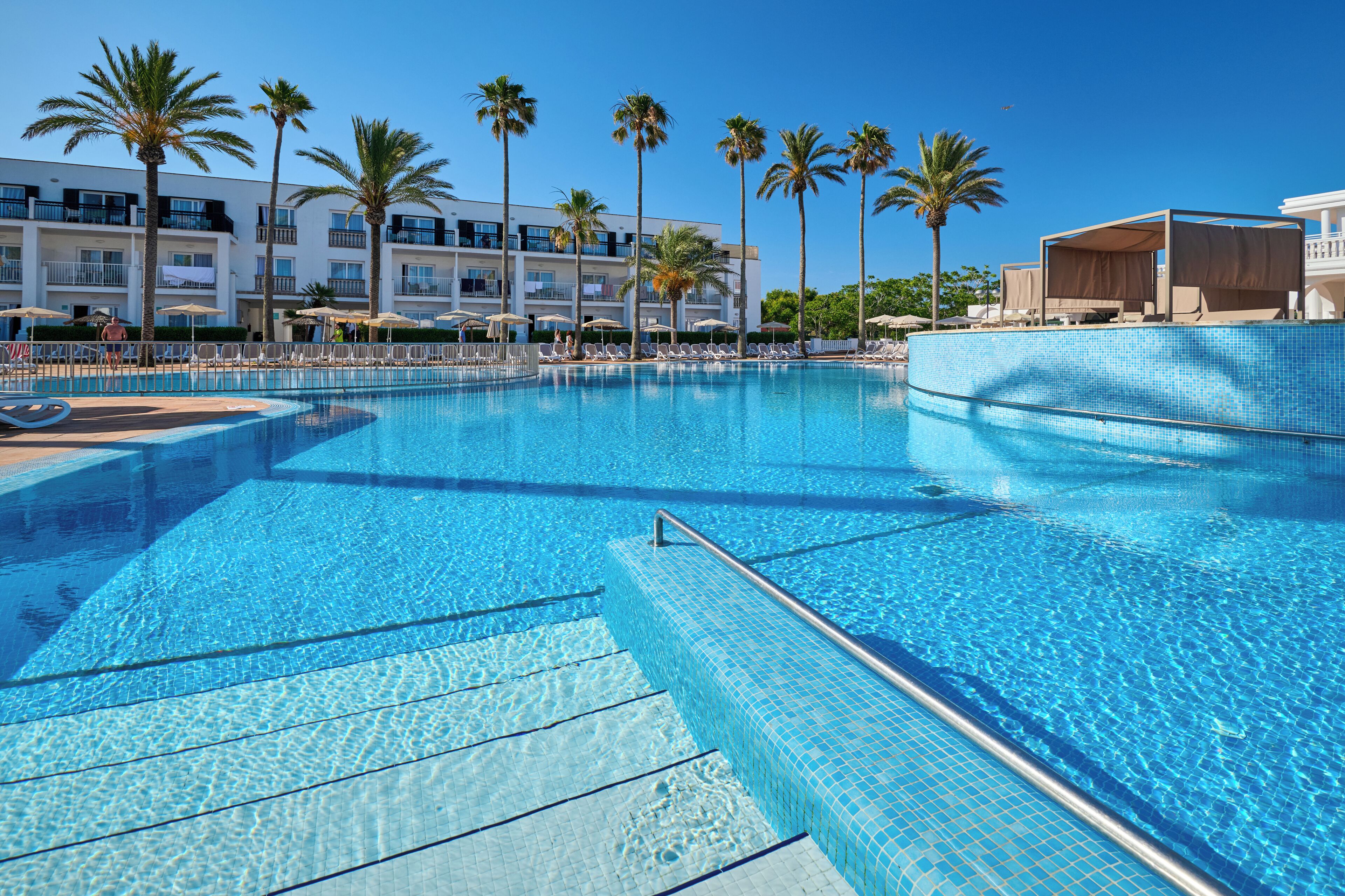 Una piscina azul cristalina con borde de azulejos, rodeada de sillones de descanso y sombrillas, con un edificio de hotel al fondo.