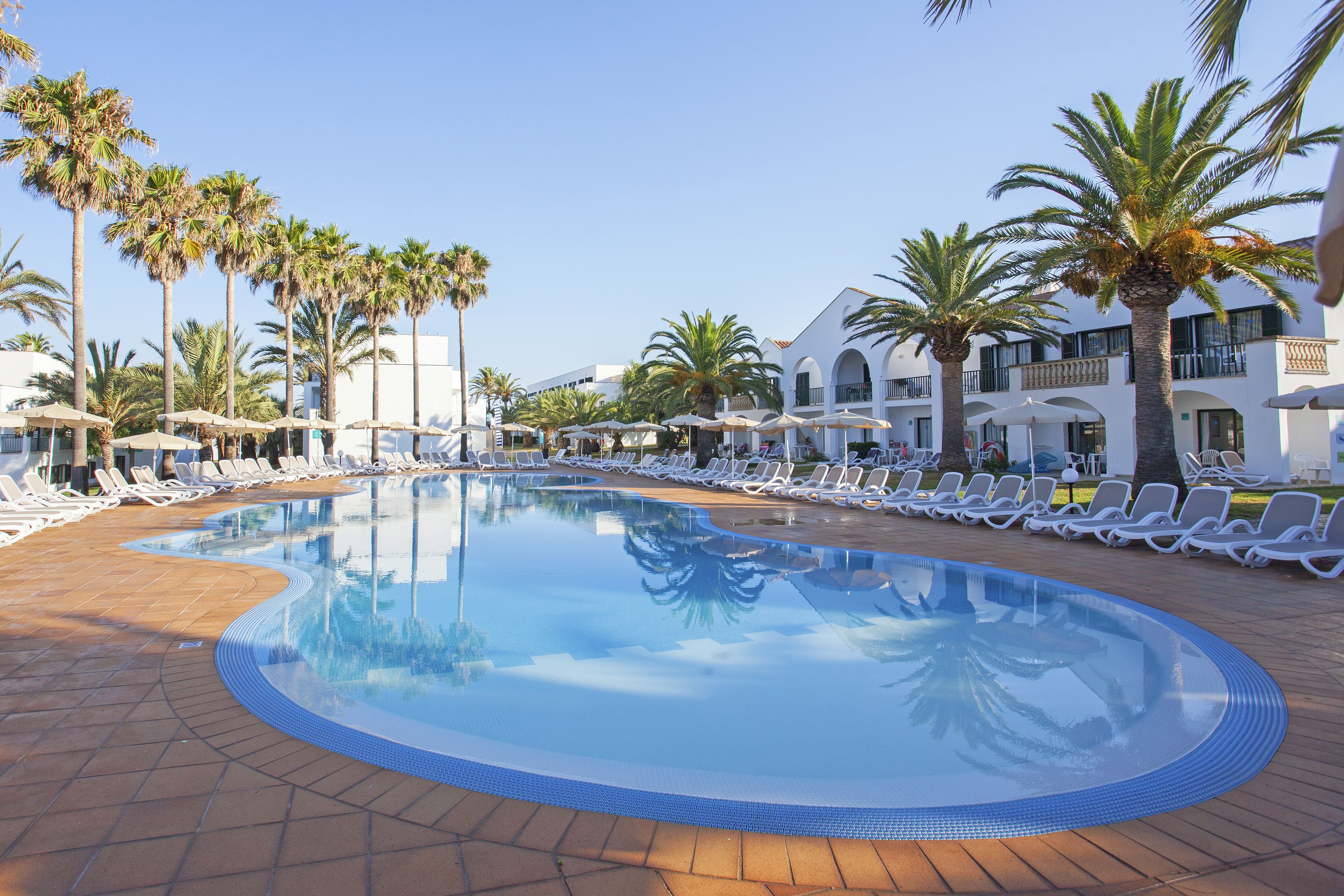 Zona de piscina con sillones reclinables y palmeras, rodeada de edificios con balcones.