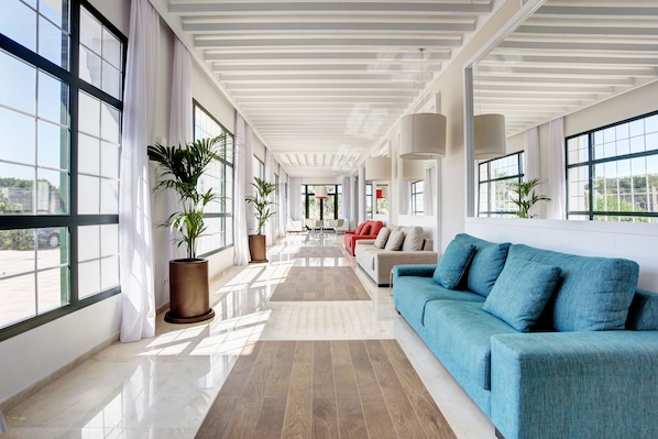 A spacious, well-lit hallway with large windows, a blue sofa, and a red chair. - Grupotel Mar de Menorca (Mahón)