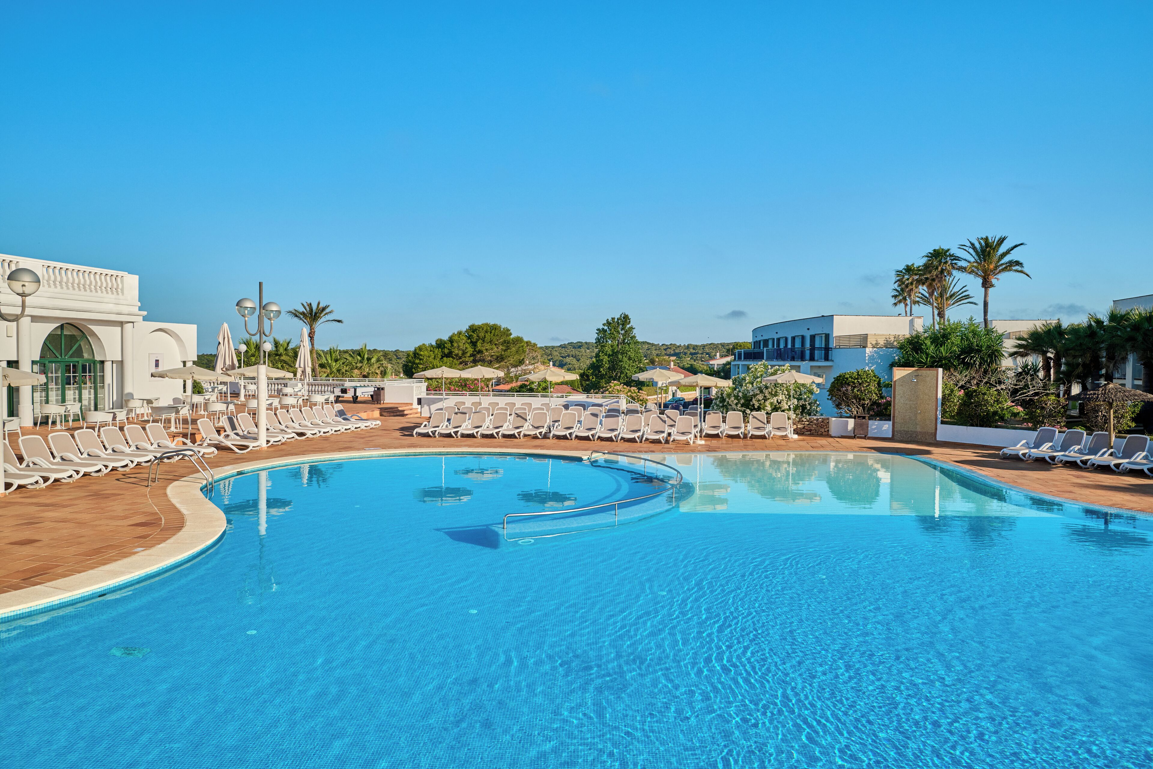 A large outdoor swimming pool with lounge chairs and umbrellas, surrounded by palm trees and buildings.