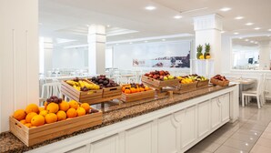 Un área de desayuno de hotel moderna con cajas de madera de frutas sobre una encimera de granito, sillas blancas y un amplio ventanal al fondo.