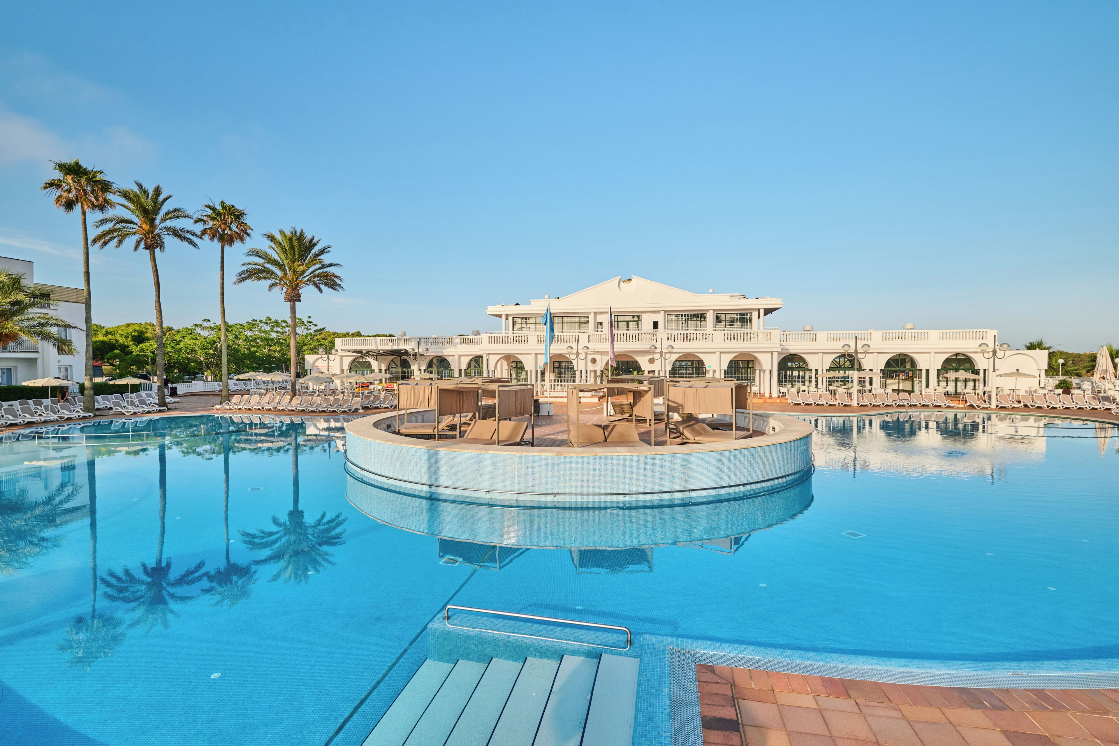 A large pool with a circular deck area, surrounded by palm trees and a building with a white facade.