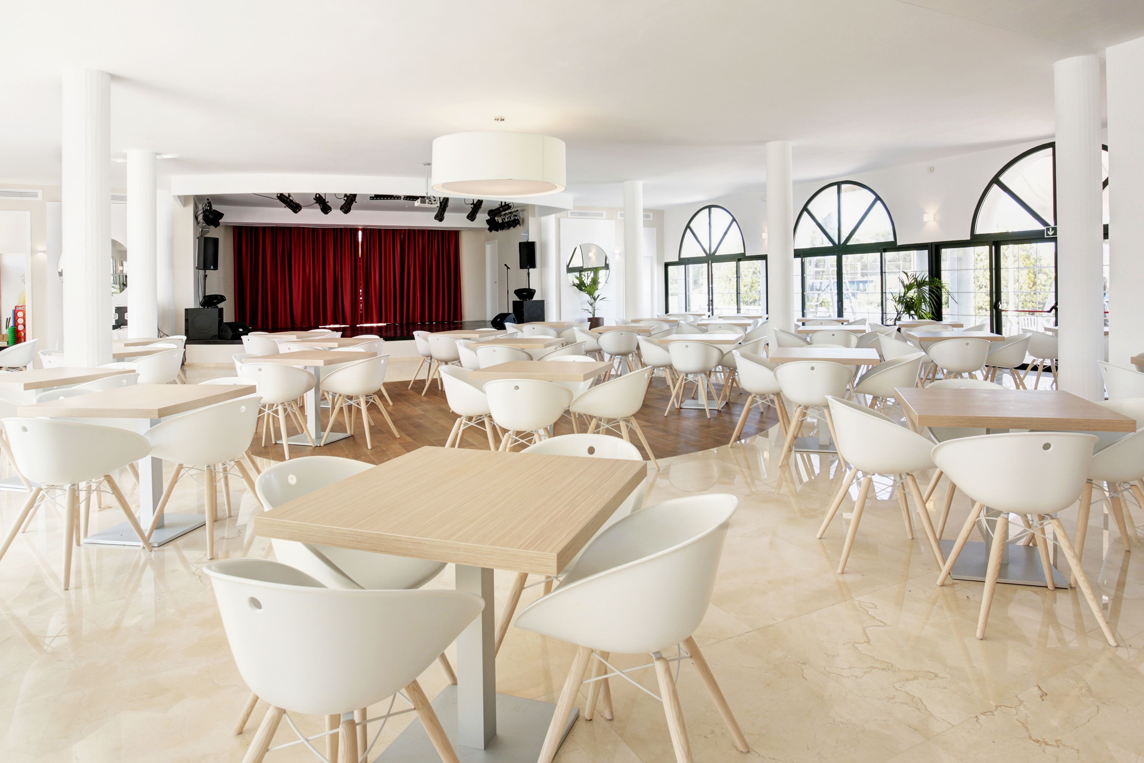 Un comedor amplio con varias mesas y sillas blancas, ventanas grandes y un escenario con cortinas rojas al fondo.