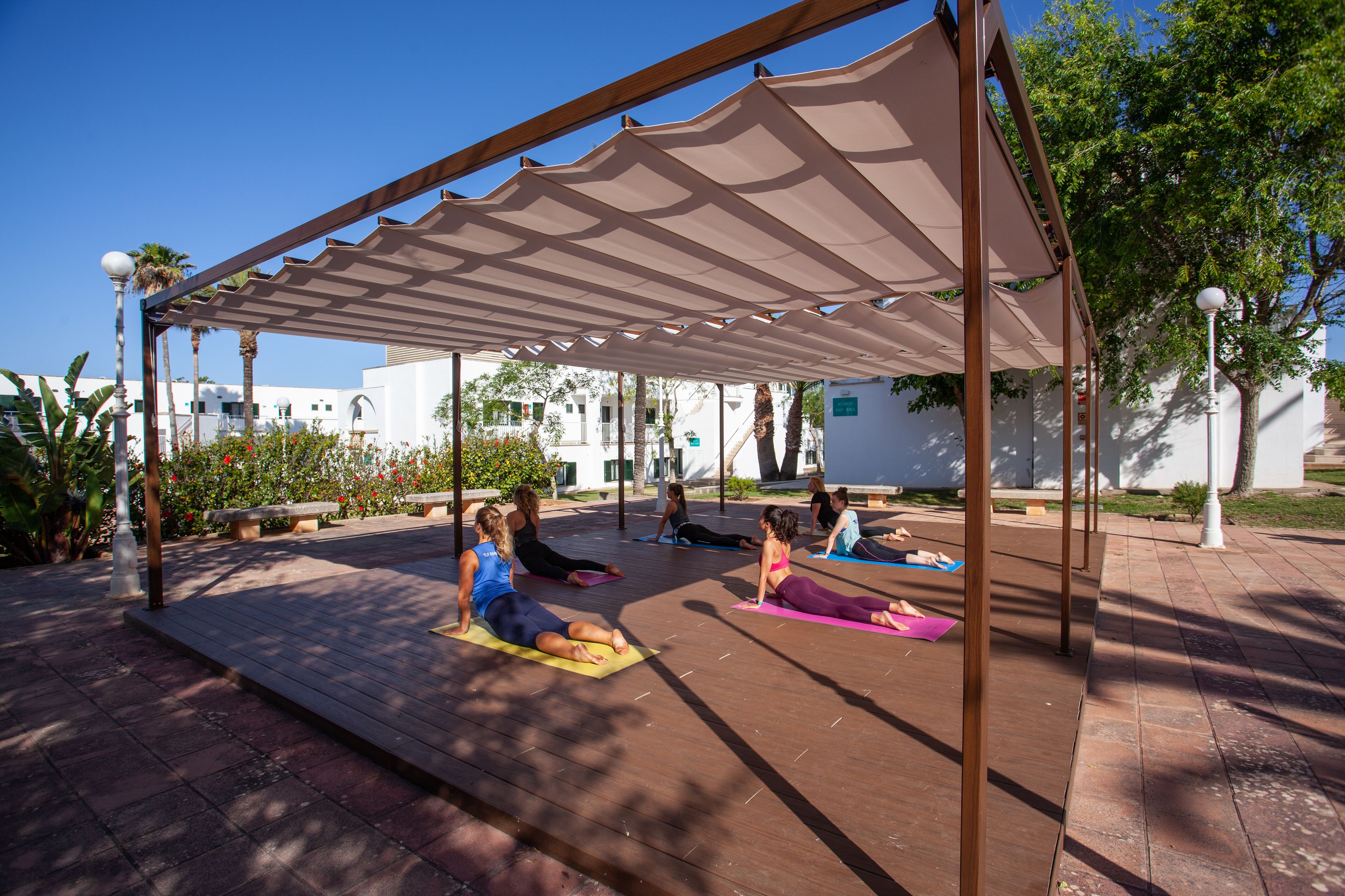 An outdoor yoga class under a shaded pavilion with a clear blue sky.