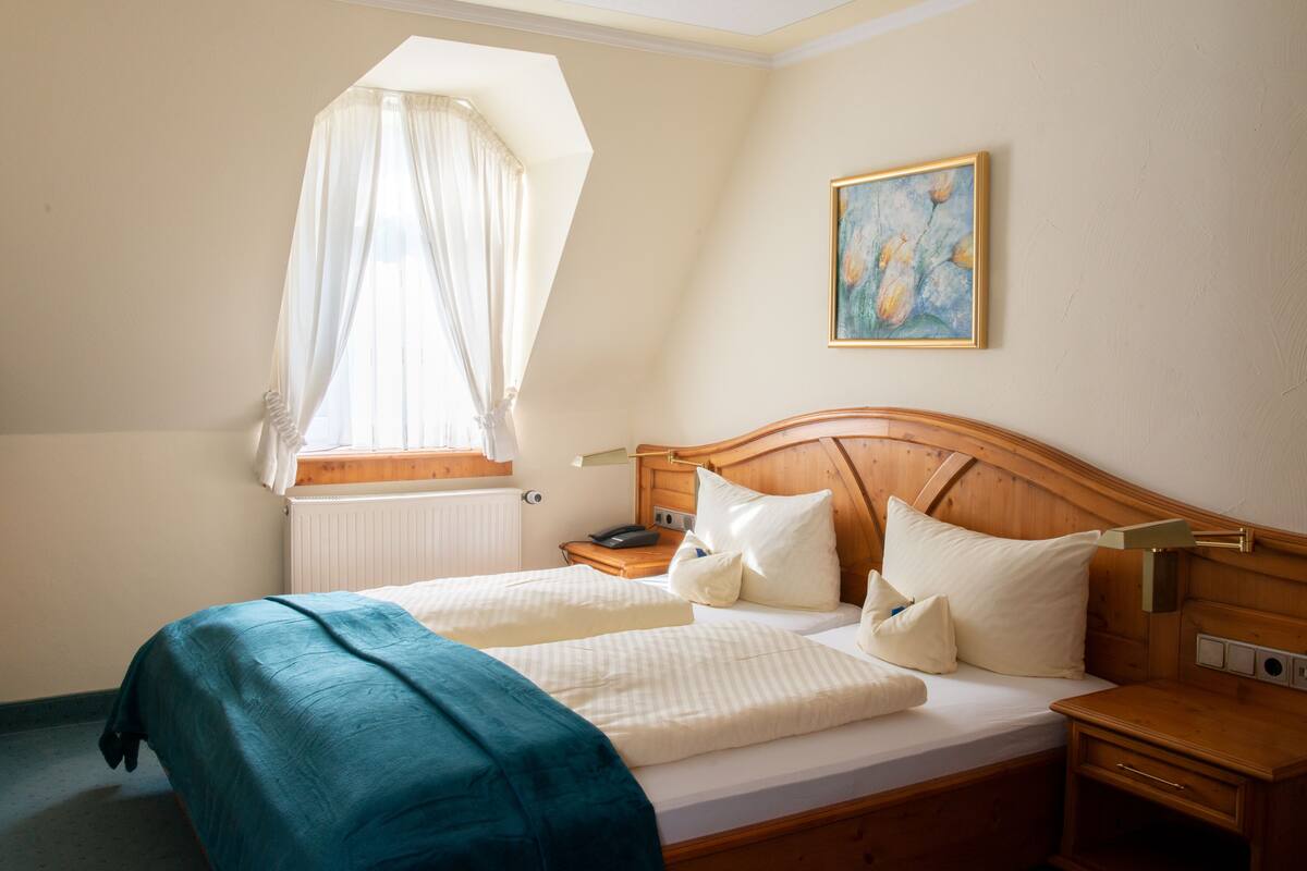 A bedroom with a wooden bed, a nightstand, a bedside table, a window with a sheer curtain, and a painting on the wall.