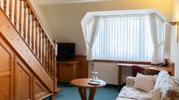 A room with a wooden staircase, a flat-screen TV, a round wooden table with a tray, a floral sofa, and a window with sheer curtains.