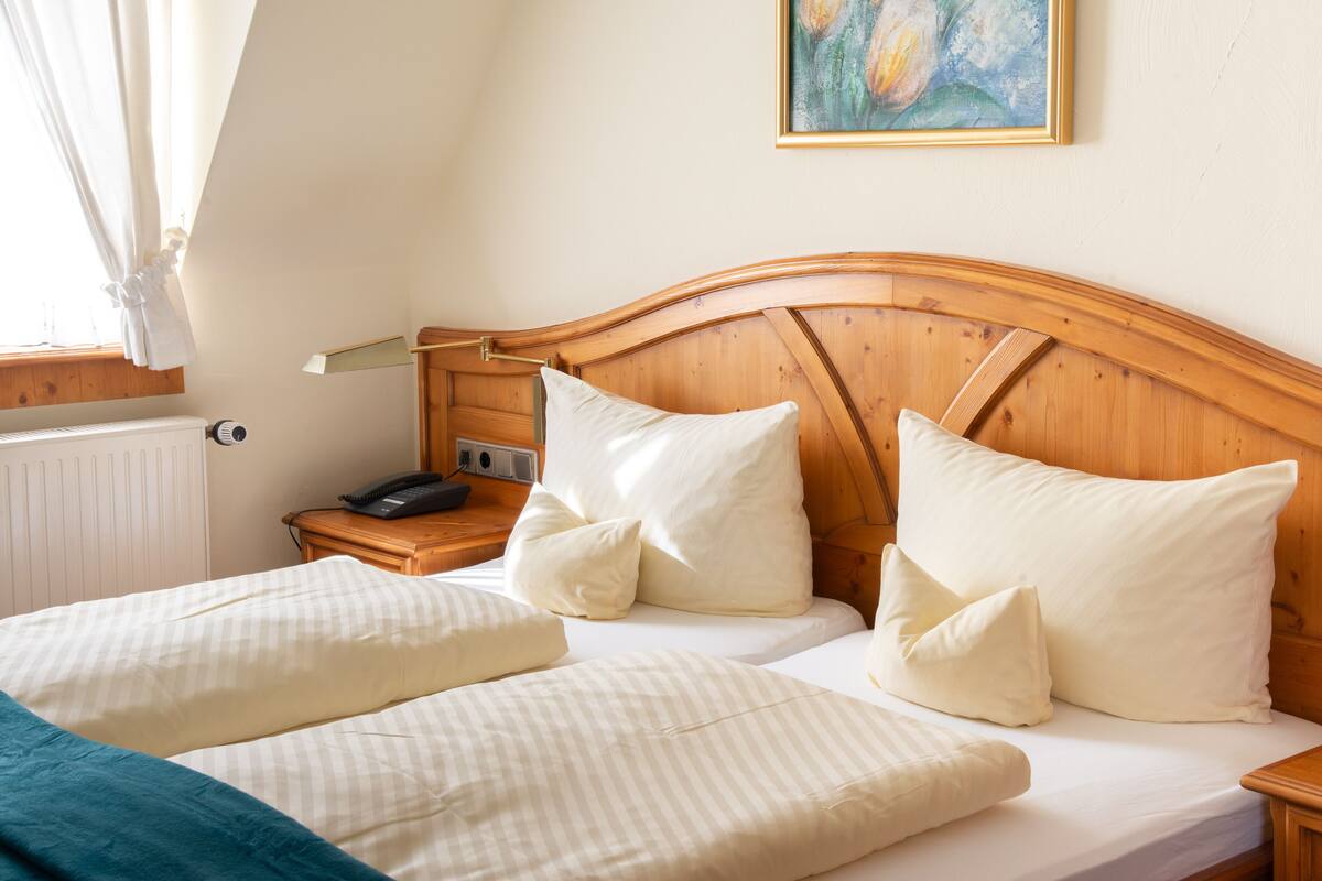 A hotel room with a wooden headboard, two beds with white bedding, a bedside table with a lamp, and a painting on the wall.