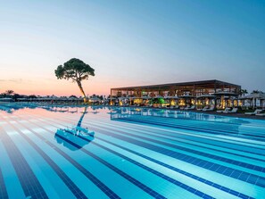 A swimming pool with lounge chairs and umbrellas, a large tree, and a building with multiple levels and outdoor seating.