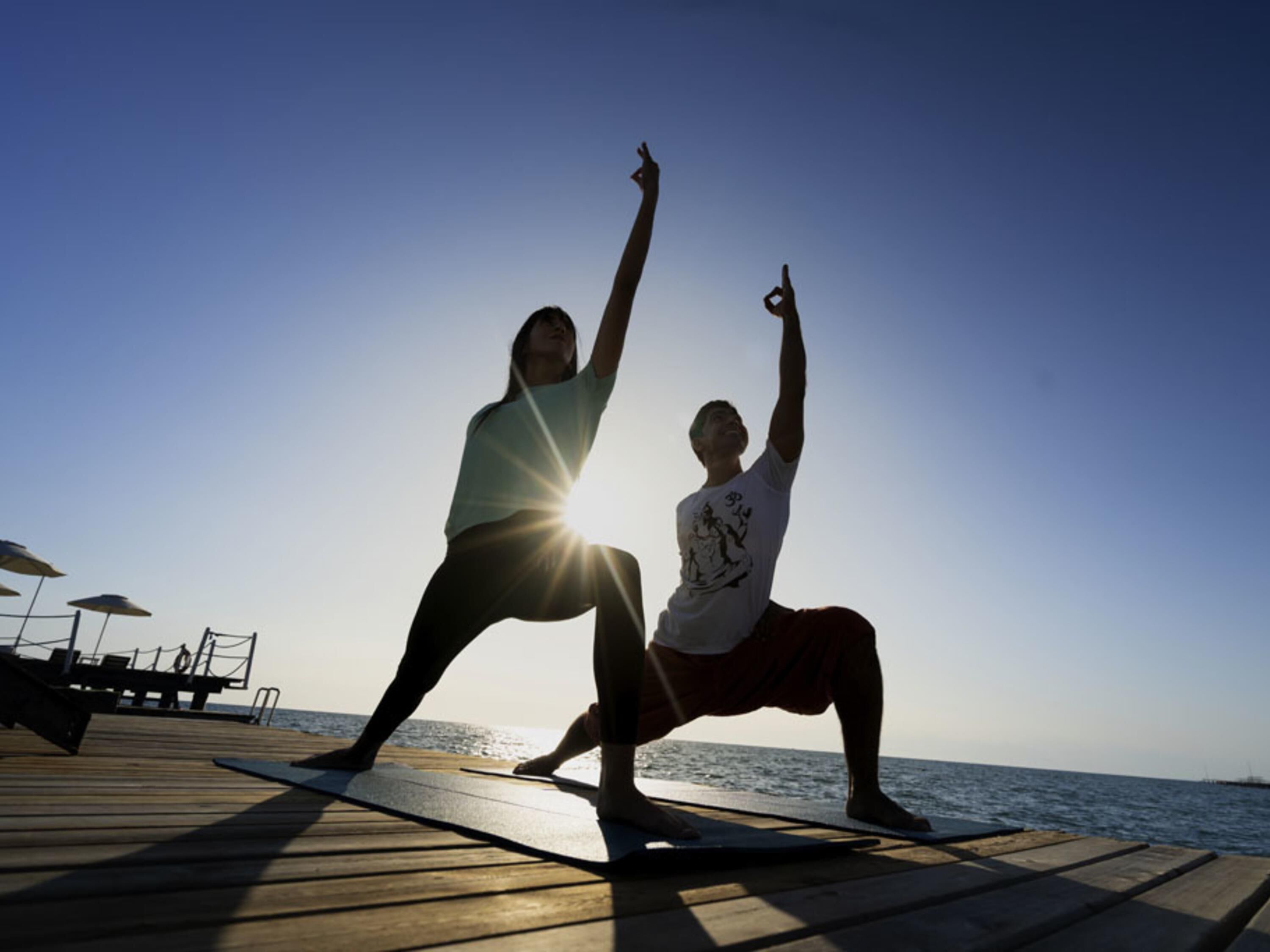 Twee personen doen yoga op een houten terras aan de zee.