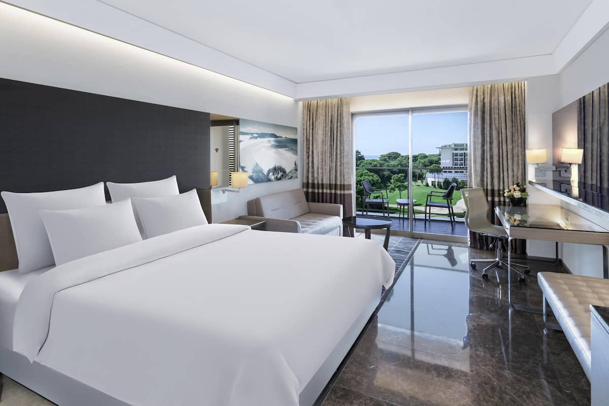 A modern hotel room with a large bed, a desk, and a balcony with a view of the city.