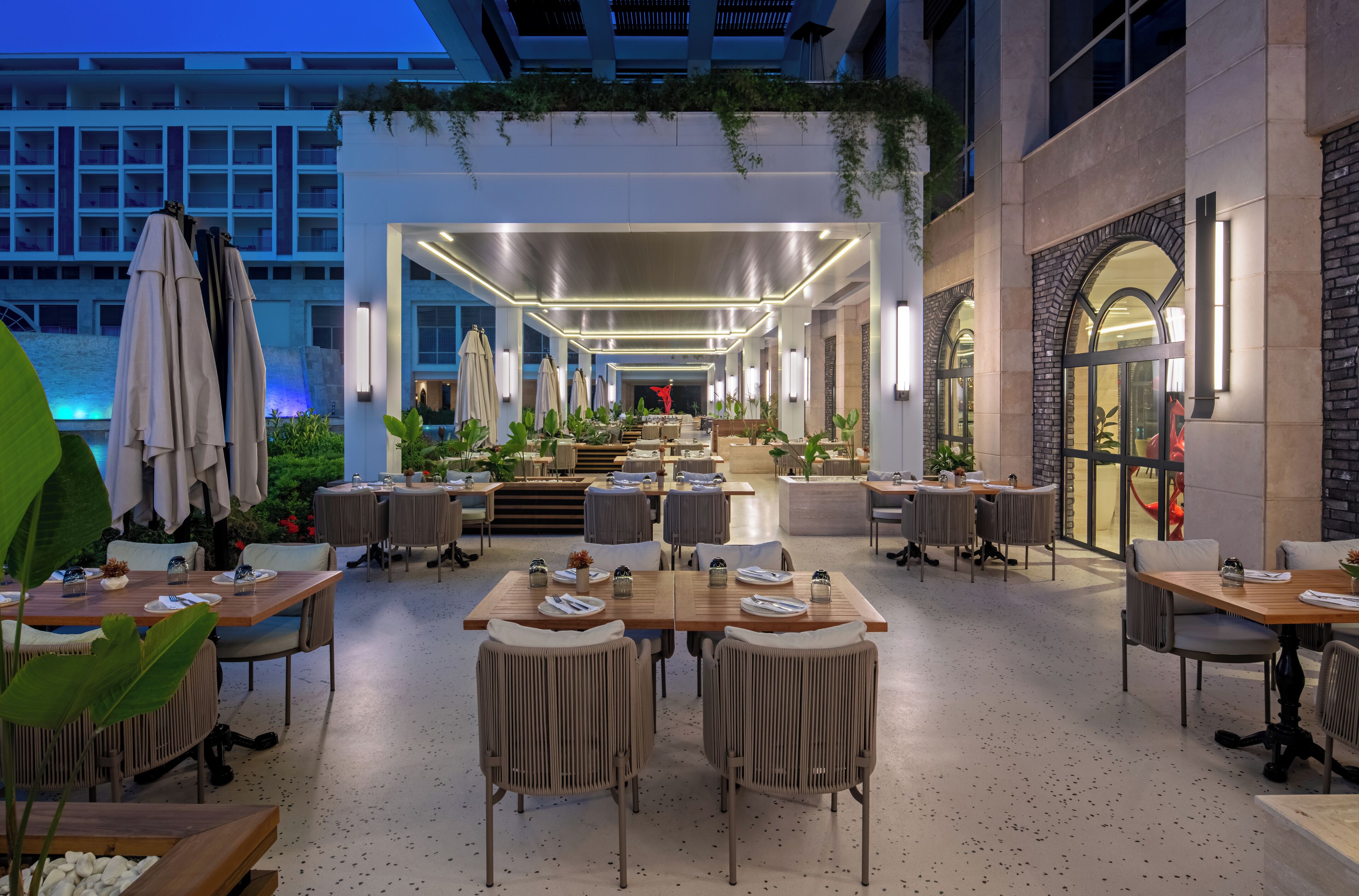 A modern outdoor dining area with multiple tables and chairs, surrounded by plants and a view of a building in the background.
