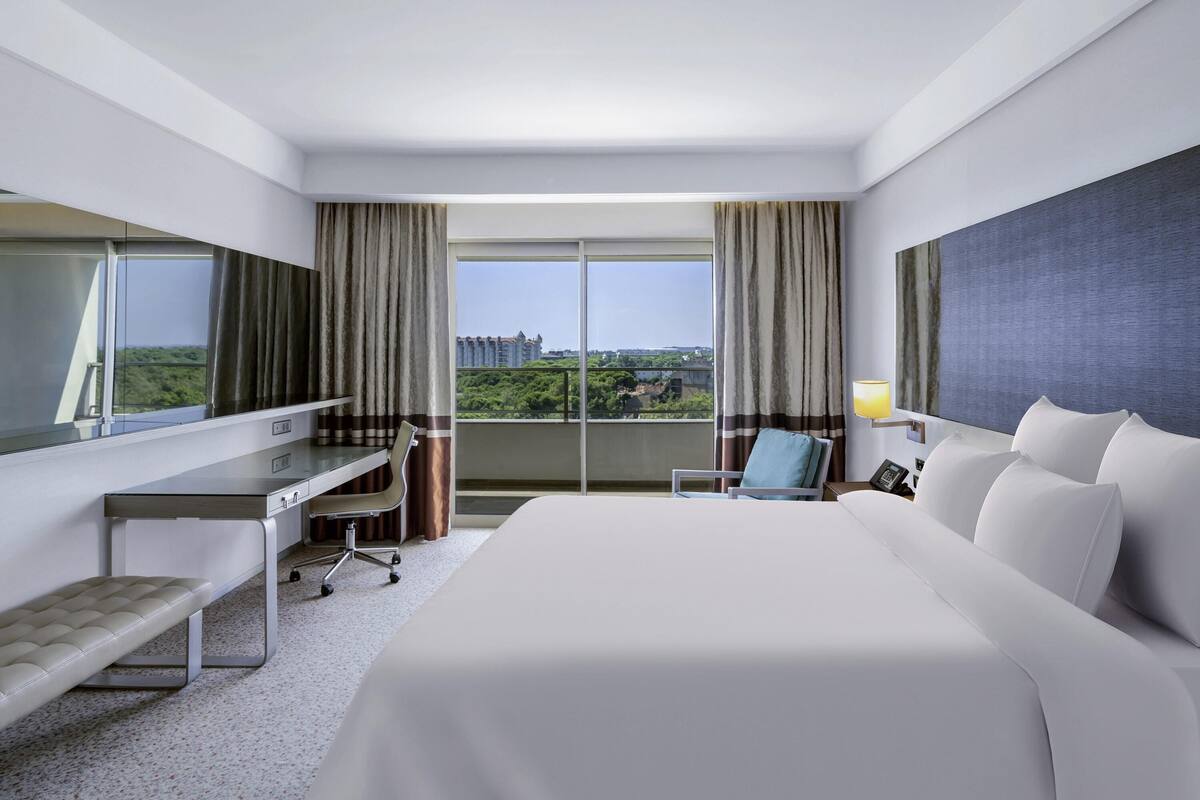 A hotel room with a bed, desk, chair, and a view of greenery and buildings.