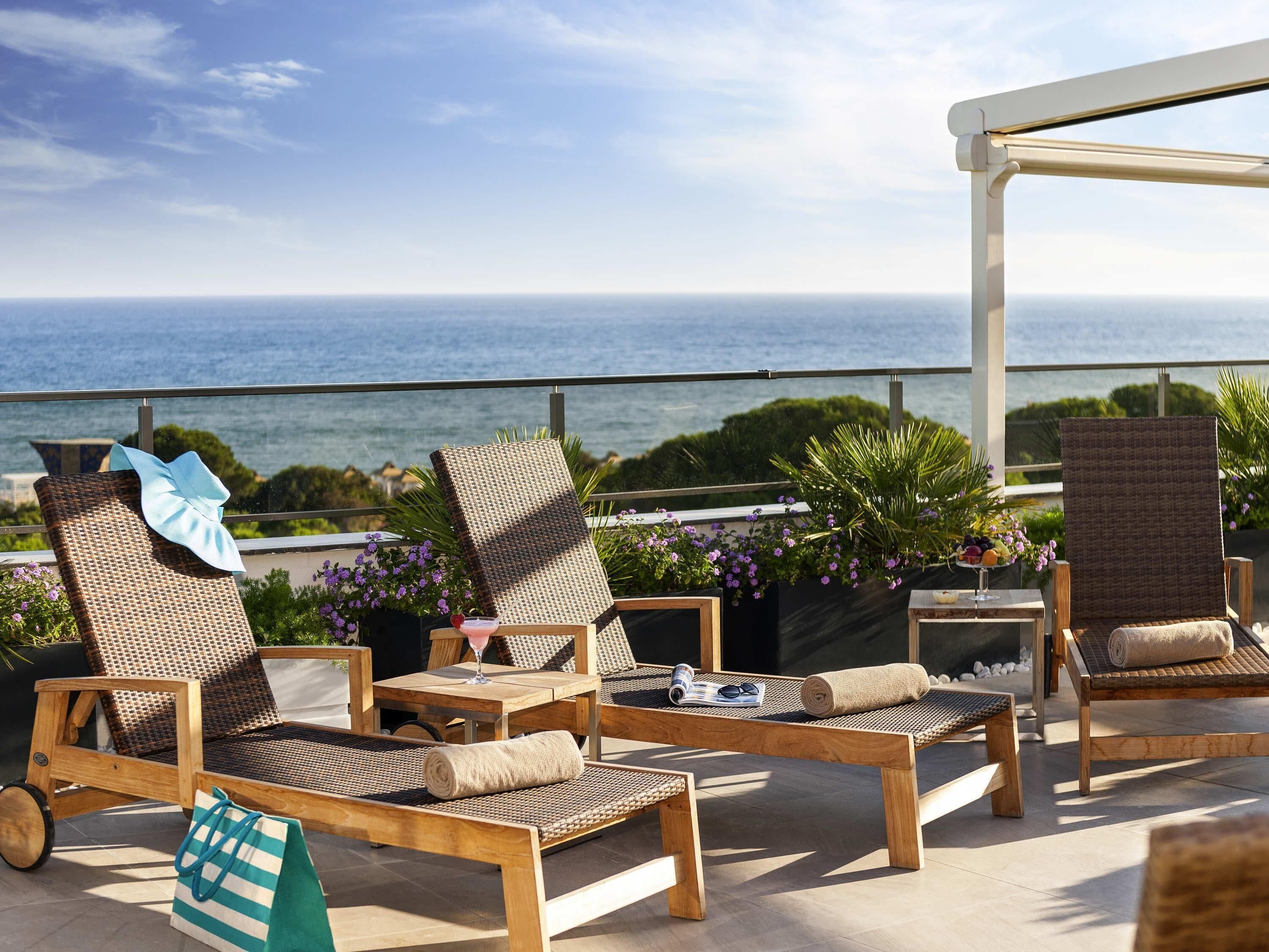 A terrace with wicker lounge chairs, a wooden table, and a view of the sea.