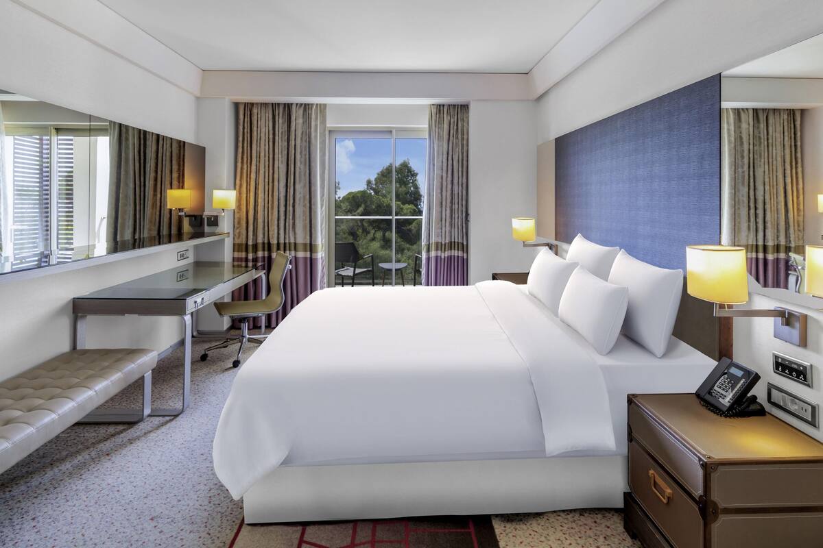 A hotel room with a large bed, a desk, a chair, a desk lamp, a bedside table with a phone, and a window with curtains.