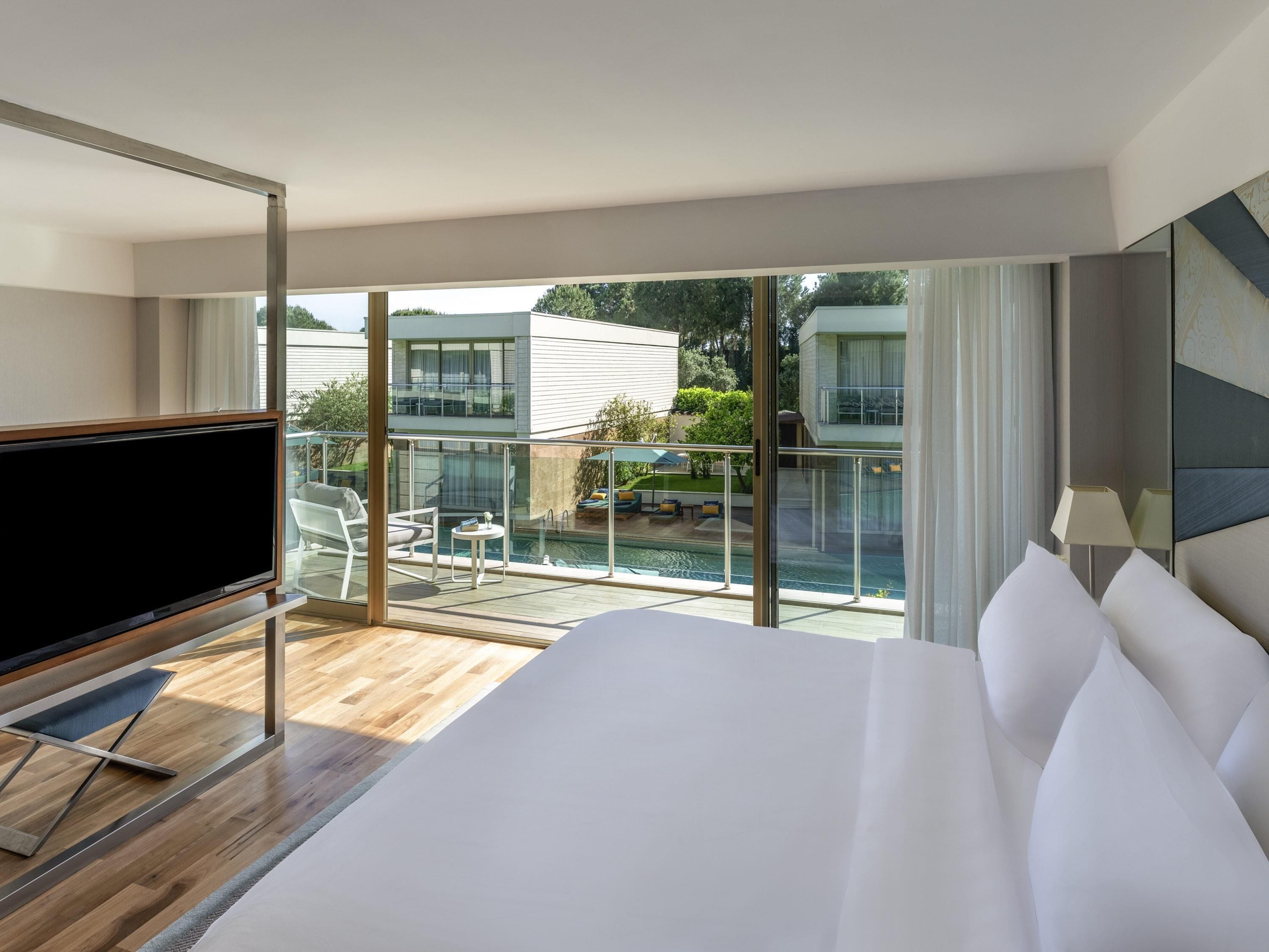 A hotel room with a large bed, a TV, a balcony with a pool, and a view of other buildings.