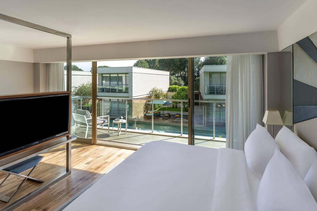 A hotel room with a large bed, a TV, a balcony with a pool, and a view of other buildings.