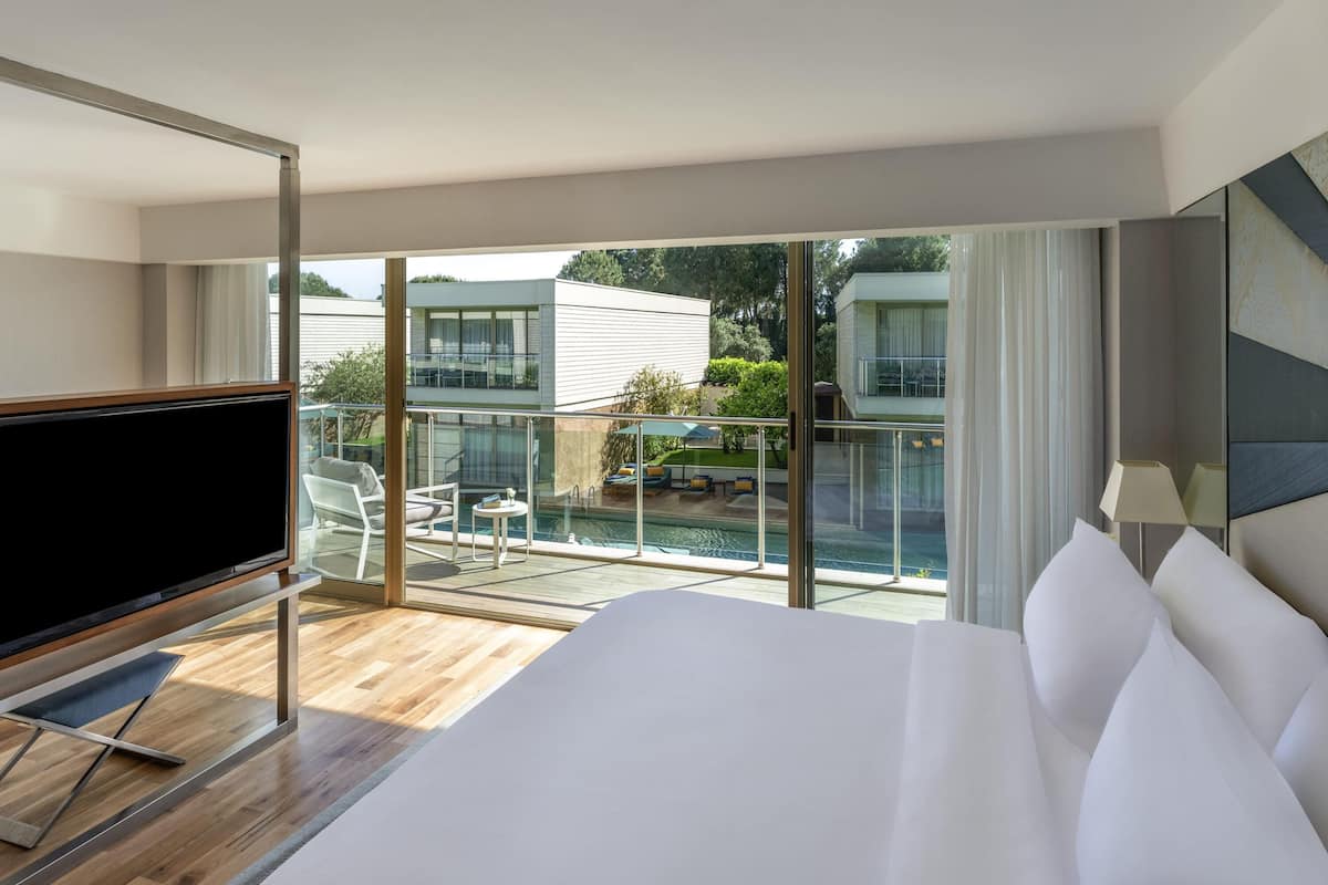 Ett hotellrum med en stor säng, en TV, en balkong med en pool och utsikt över andra byggnader.