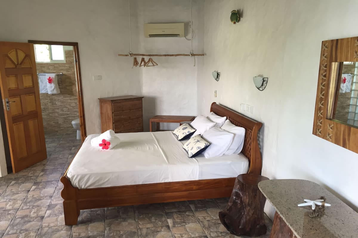 A bedroom with a wooden bed, stone-tiled floor, a wooden dresser, and a mirror with a wooden frame.