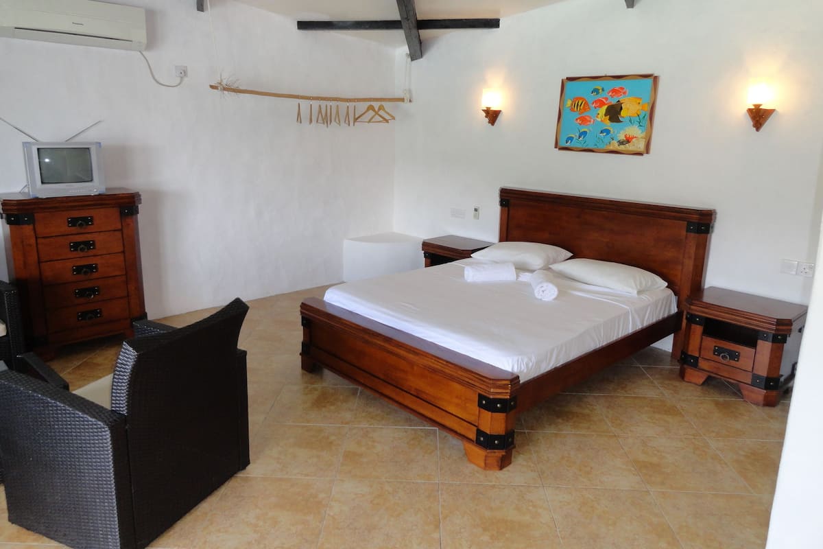 A bedroom with a wooden bed, a small bedside table, a chair, a dresser, a television, and a wall-mounted light fixture.