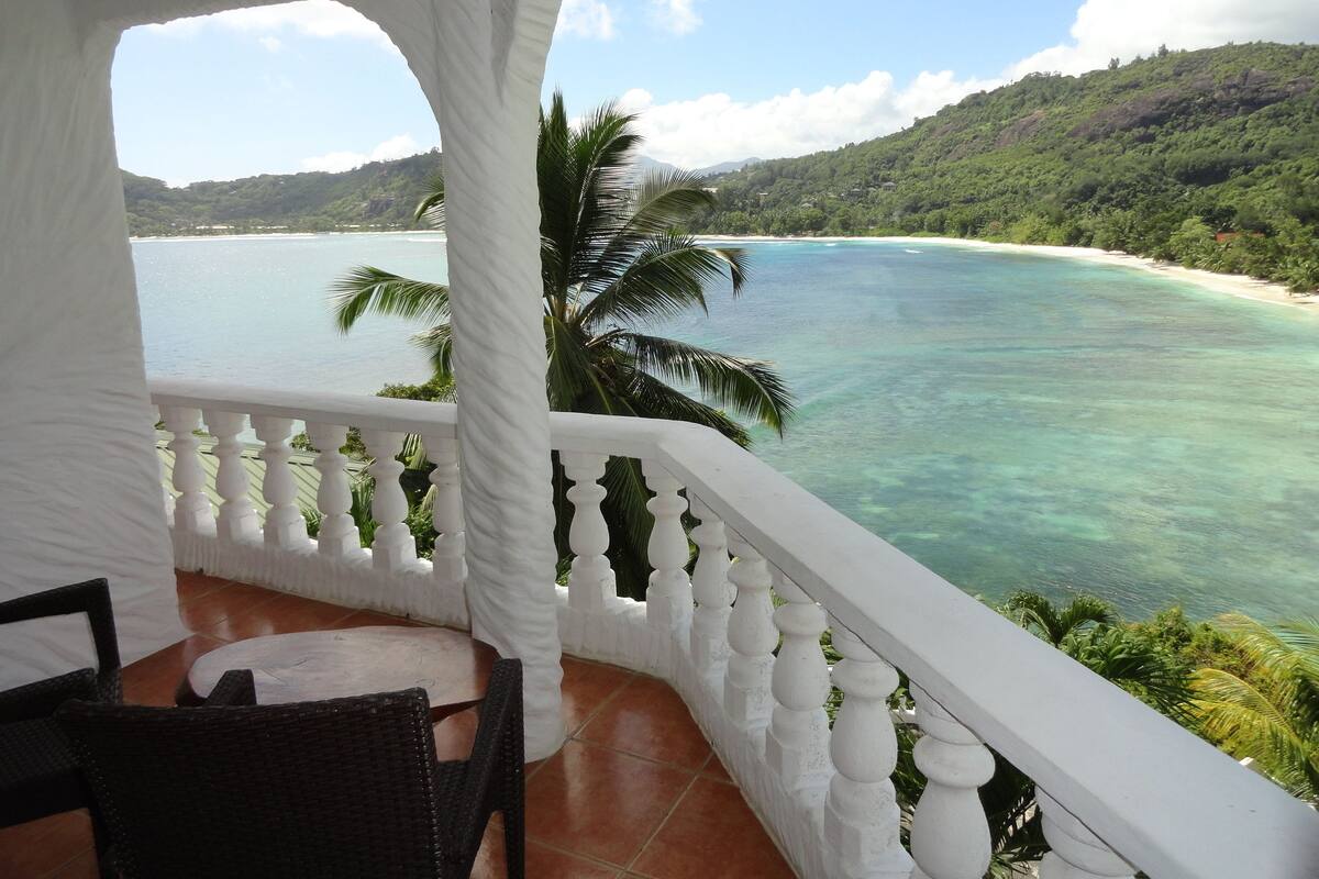 A balcony with a white balustrade overlooking a turquoise sea and a lush green landscape.