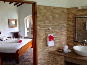 A hotel room with a wooden bed, a stone-tiled floor, a stone wall, a bathroom with a sink and mirror, and a towel rack with a towel and a red flower. - Lazare Picault Hotel (Mahé Island)