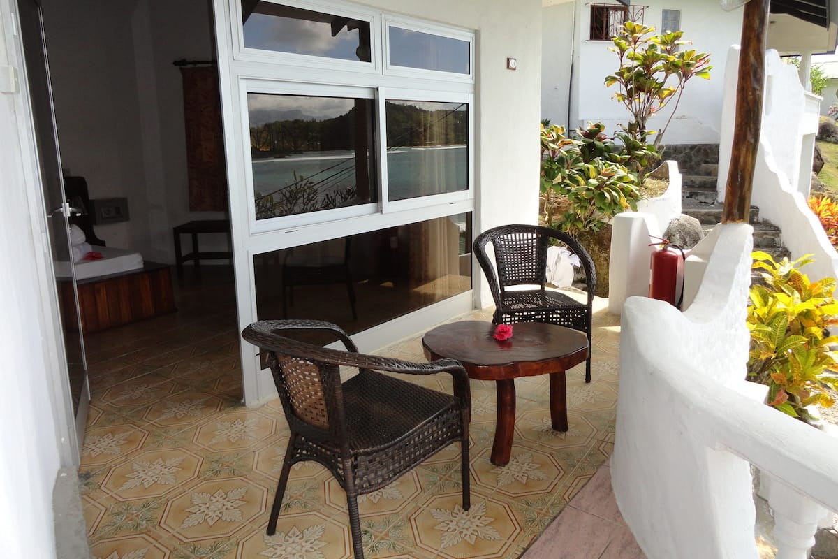 A patio with two wicker chairs and a small table, a white wall, and a view of a mountainous landscape through a window.
