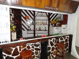 Una zona de bar con una recepción, taburetes de madera, una pared de piedra y estanterías con botellas y vasos.