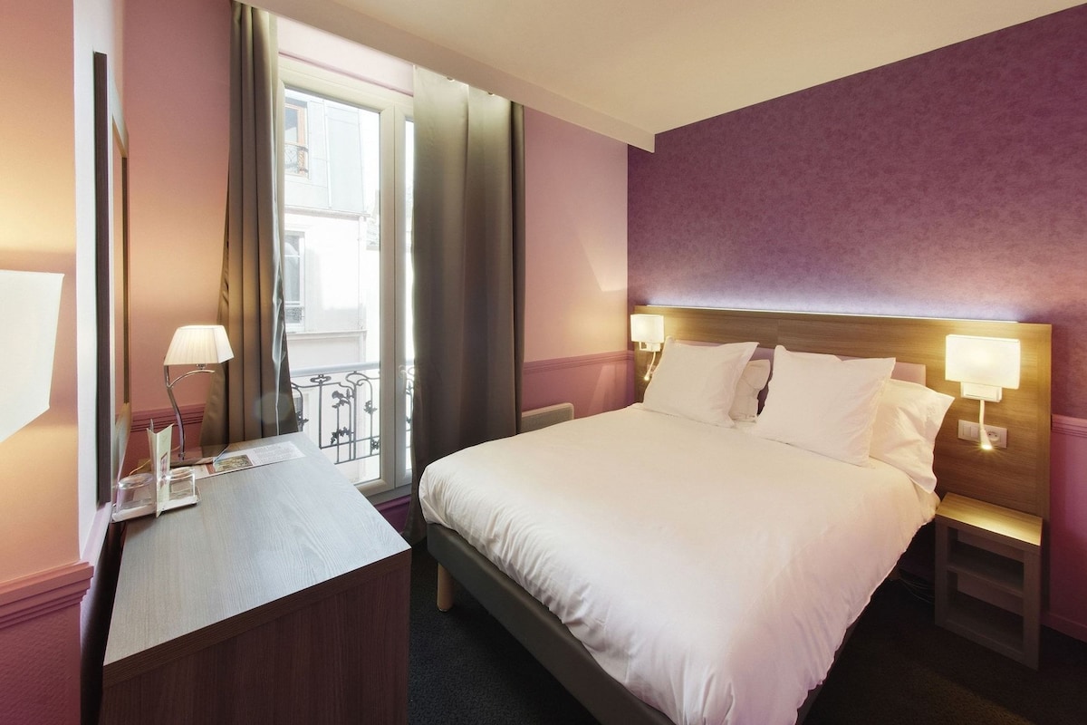 A hotel room with a bed, bedside tables, a desk with a lamp, and a window with curtains.