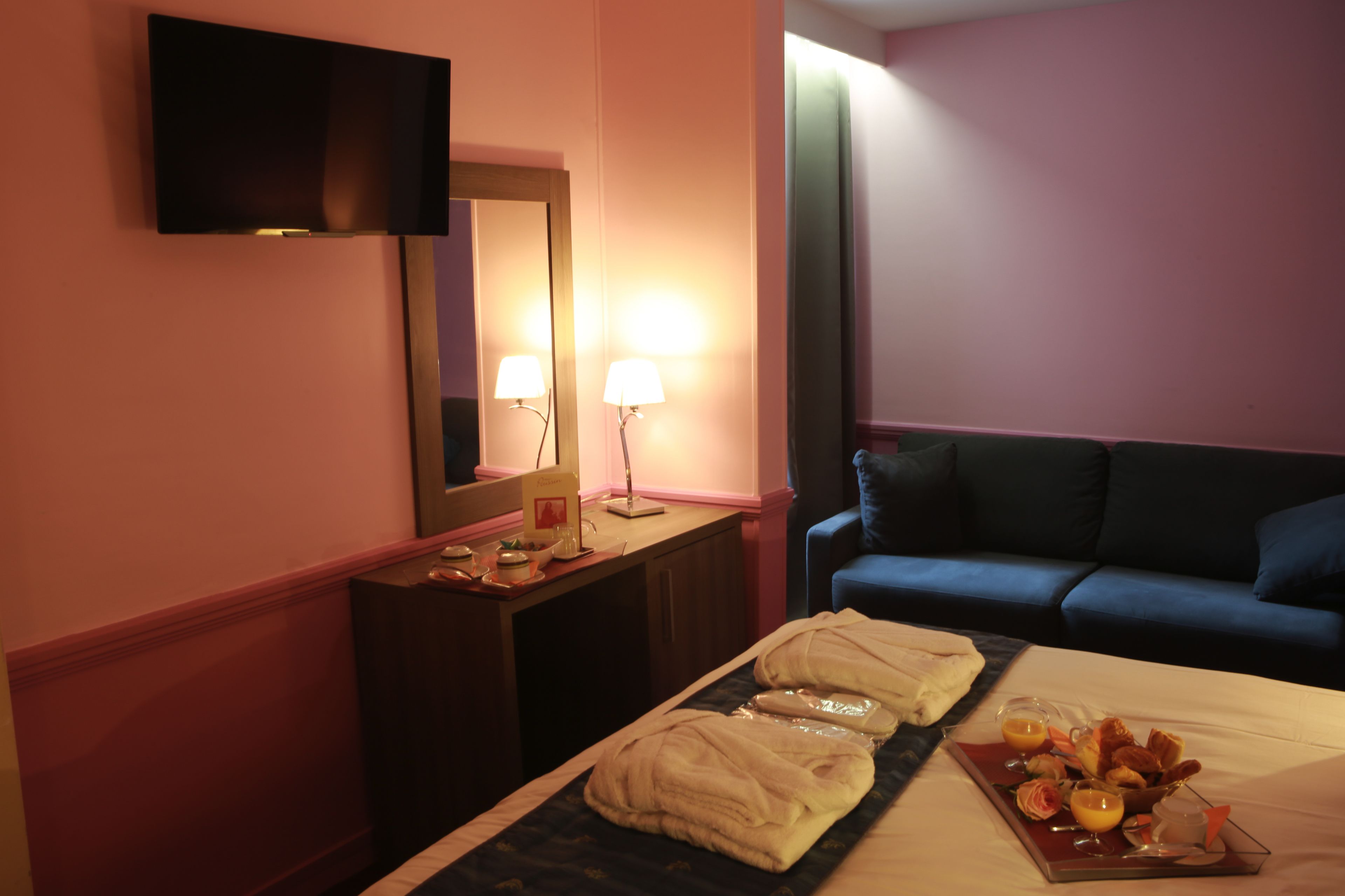 Una habitación de hotel con cama, una mesita de noche con lámpara, un televisor de pantalla plana montado en la pared y un sofá azul.