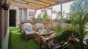 Un porche cubierto con mobiliario de mimbre, una mesa de vidrio y un pequeño jardín con plantas en macetas.