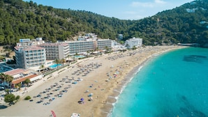 Un hotel frente al mar con varias construcciones, una playa y montañas al fondo.