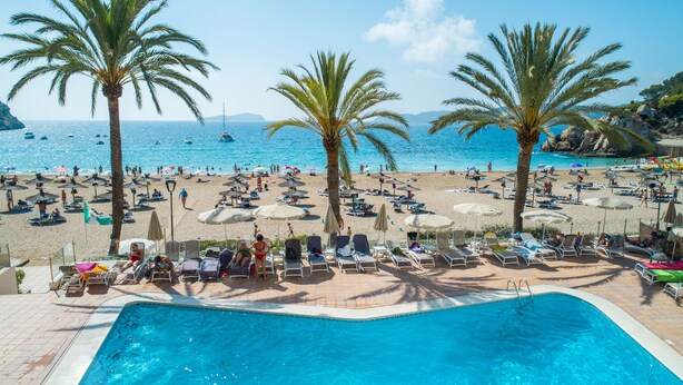 Vista de la piscina de un resort frente a la playa, con tumbonas, sombrillas y palmeras.