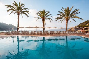 A pool with lounge chairs and umbrellas, palm trees, and a view of the sea.